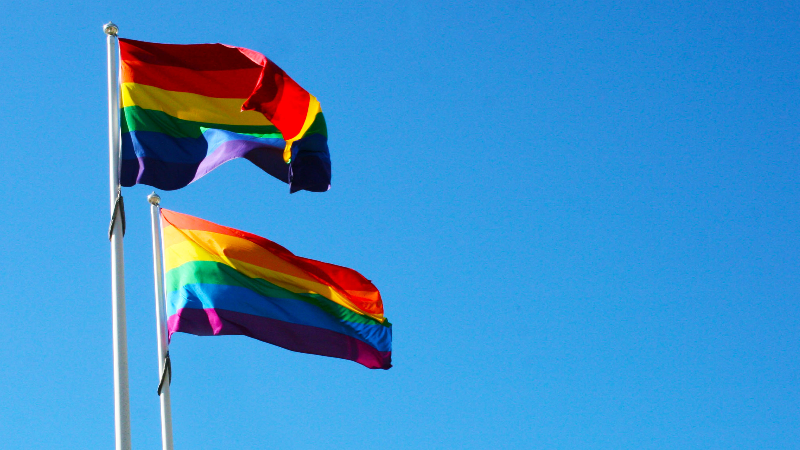 Två prideflaggor vajar i vinden mot en blå himmel.